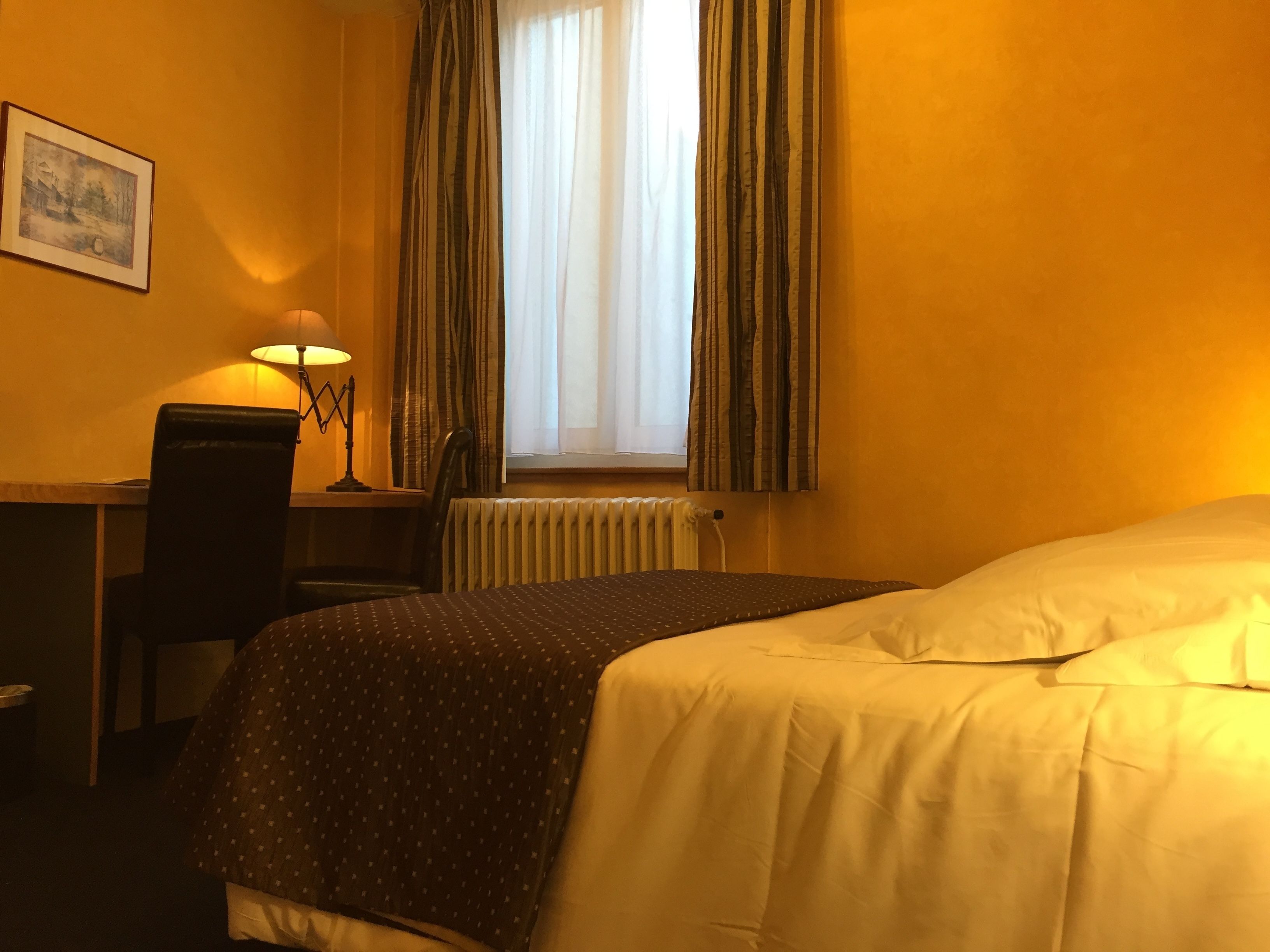 A hotel room with a bed, a desk with a lamp, a chair, a radiator, and a window with curtains.
