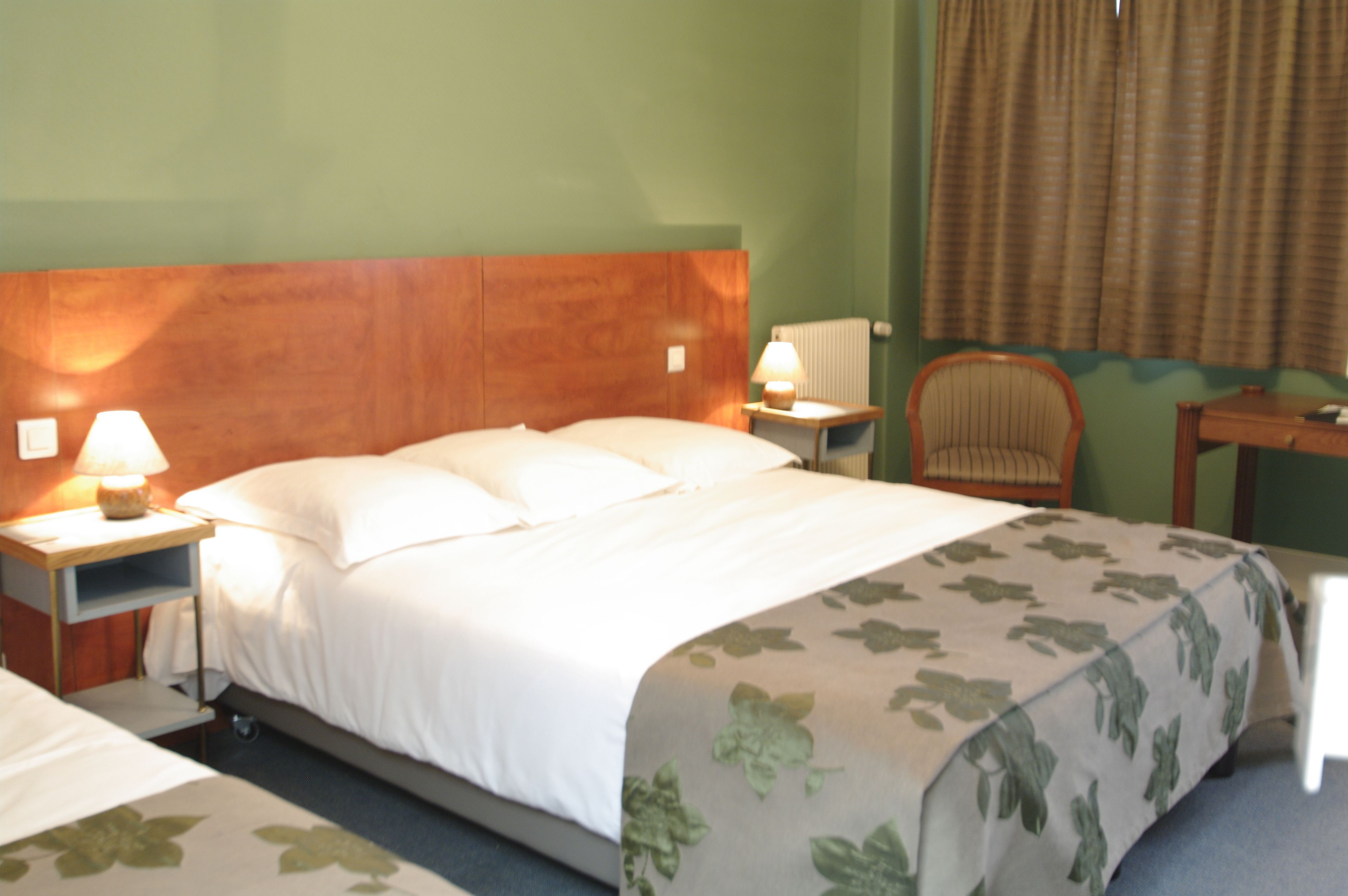 Habitación de hotel con cabecera de madera, cama con ropa blanca, mesita de noche con lámpara, una silla y una pequeña mesa.
