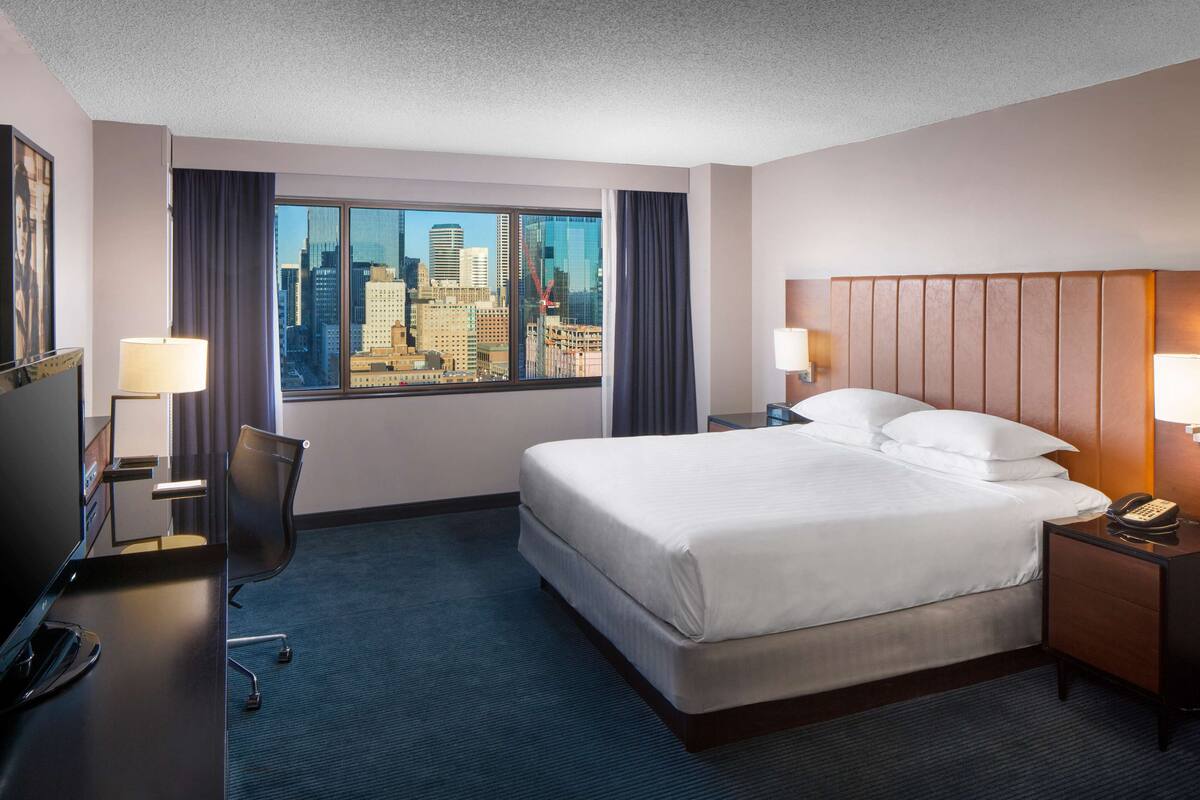 A hotel room with a large bed, two bedside tables with lamps, a desk with a chair, and a view of a cityscape through a large window.