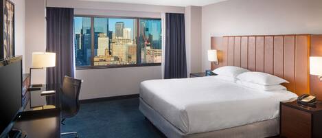 Habitación de hotel con una cama grande, dos mesitas de noche con lámparas, un escritorio con silla y vistas a un paisaje urbano a través de un ventanal amplio.