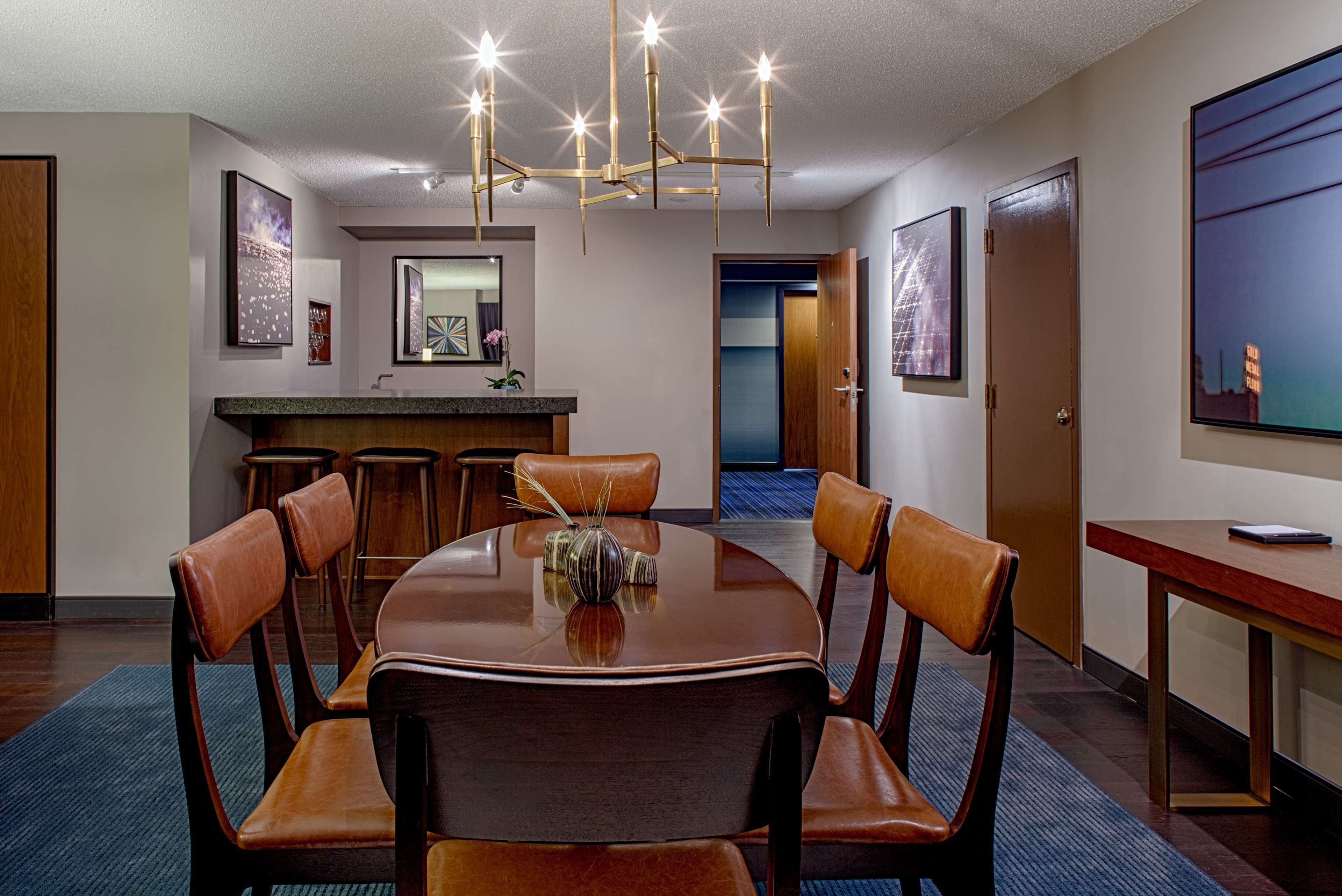 A conference room with a round table, six chairs, a chandelier, a bar area, and framed artwork on the walls.