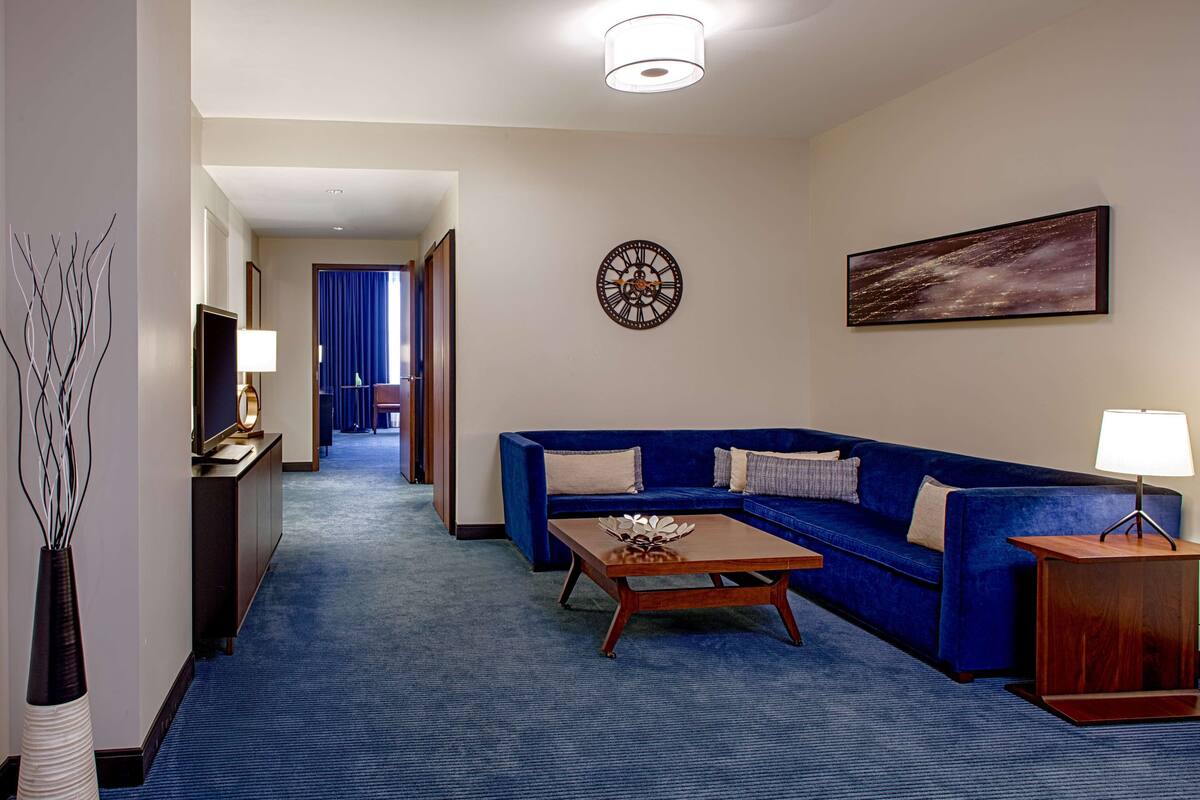 A hotel room with a blue carpet, a sofa, a coffee table, a TV, a clock, and a painting on the wall.
