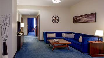 Ein Hotelzimmer mit blauem Teppich, einer Couch, einem Couchtisch, einem Fernseher, einer Uhr und einem Bild an der Wand.