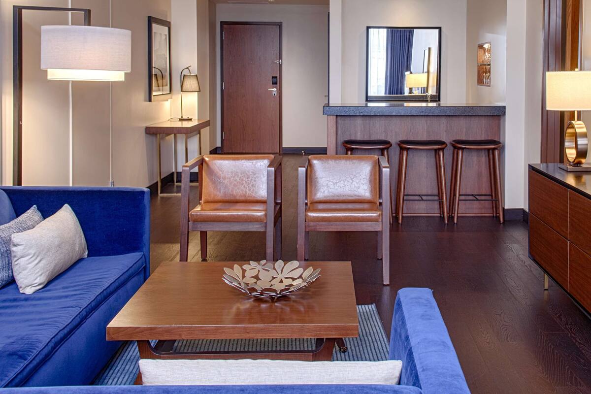 A modern hotel room with a blue sofa, two leather chairs, a wooden coffee table with a decorative centerpiece, a kitchen area with a bar stool, and a television on a wooden cabinet.