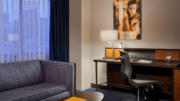 A hotel room with a sofa, a desk with a chair, a lamp, and a painting on the wall.
