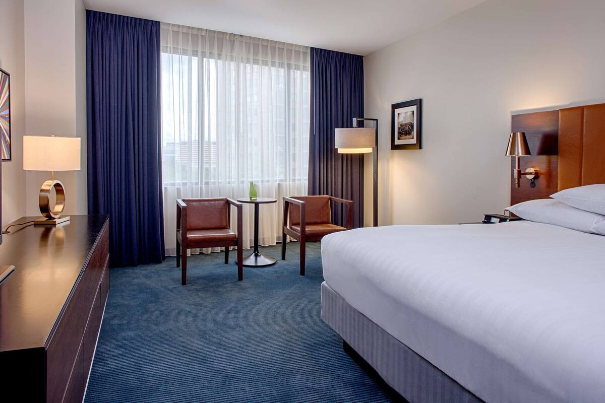 A hotel room with a large bed, a desk with a flat-screen TV, two chairs, a small table, and a window with curtains.