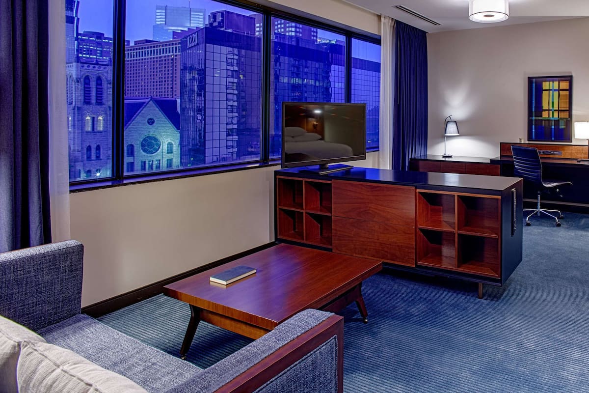 A hotel room with a large window, a desk, a television, a sofa, and a chair.