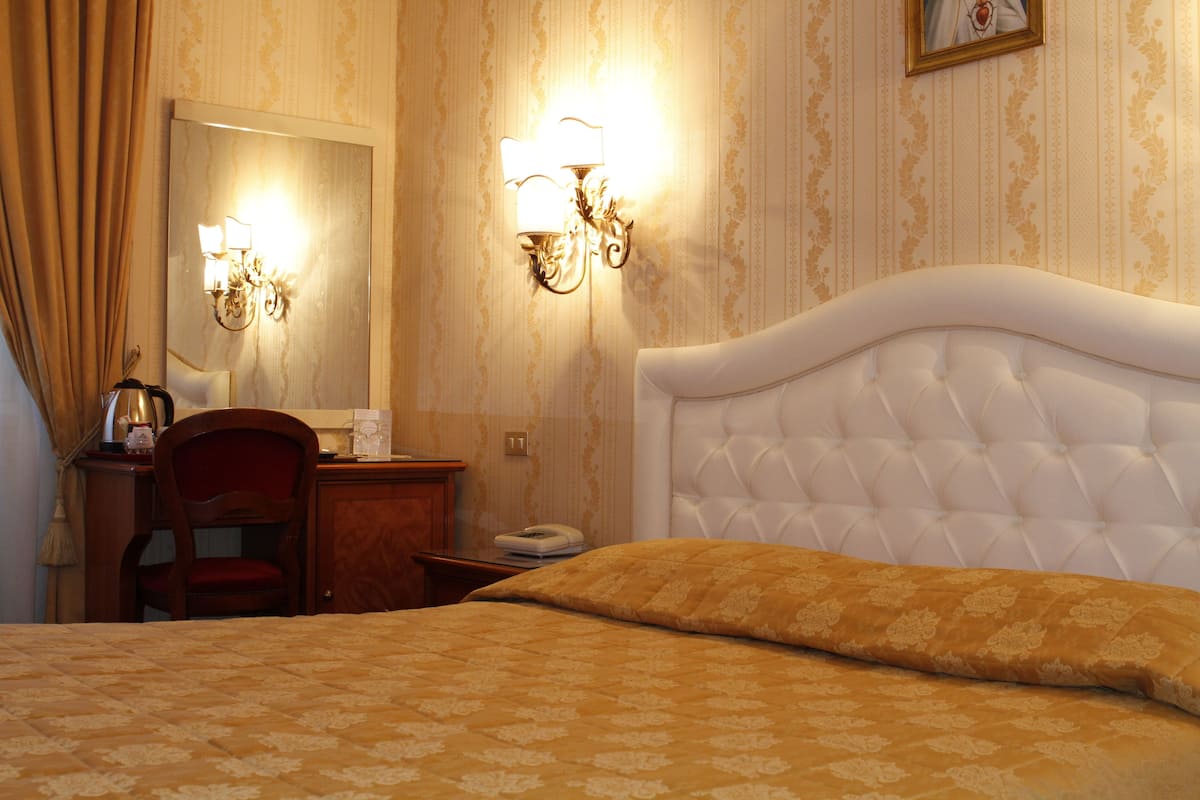 A hotel room with a large bed, a wooden nightstand, a chair, and a wall-mounted lamp.
