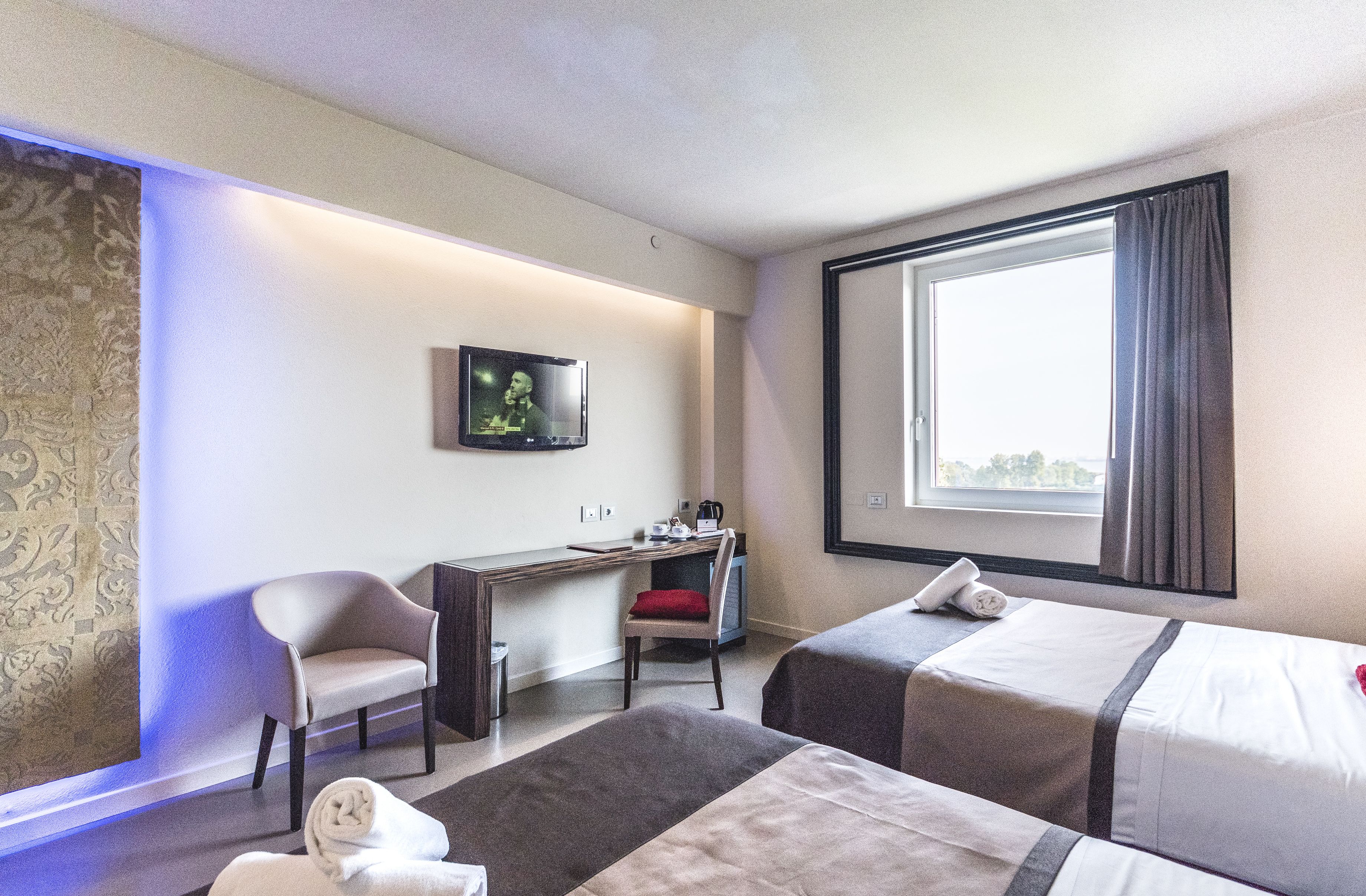 A hotel room with two beds, a desk, a chair, a TV, and a large window.