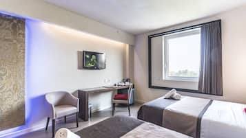 Quarto de hotel com duas camas, uma mesa, uma cadeira, uma TV e uma janela grande.