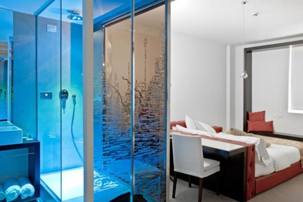 Una habitación de hotel moderna con un separador de vidrio, una cama con ropa de cama roja y blanca, un escritorio con una silla y un espejo grande en la pared.