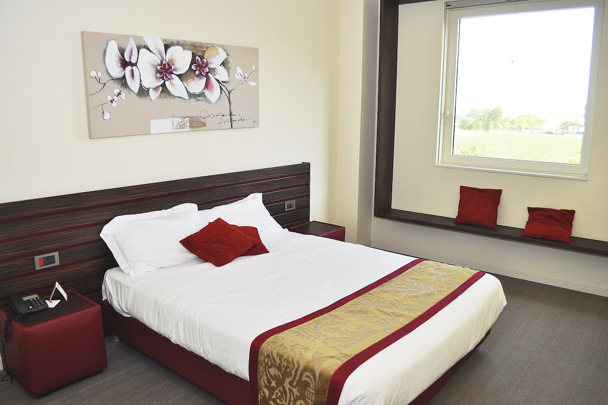 A hotel room with a large bed, a bedside table with a phone, a red pillow, and a window with a view of the outdoors.