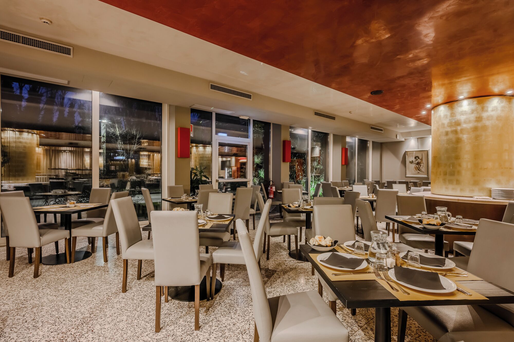 Un restaurante moderno con varias mesas, asientos cómodos y grandes ventanales que ofrecen vistas del interior.
