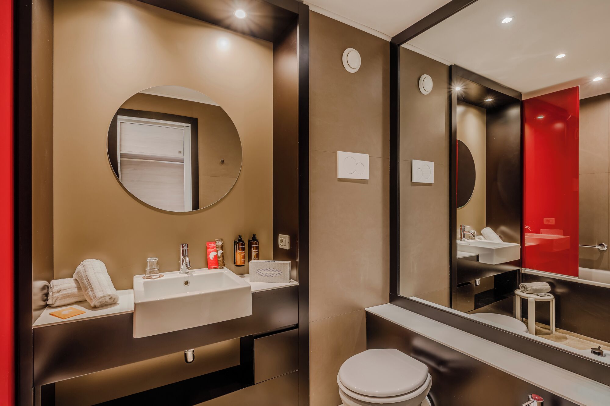 A modern bathroom with a round mirror, a white sink, and a toilet. There are toiletries on the sink counter.