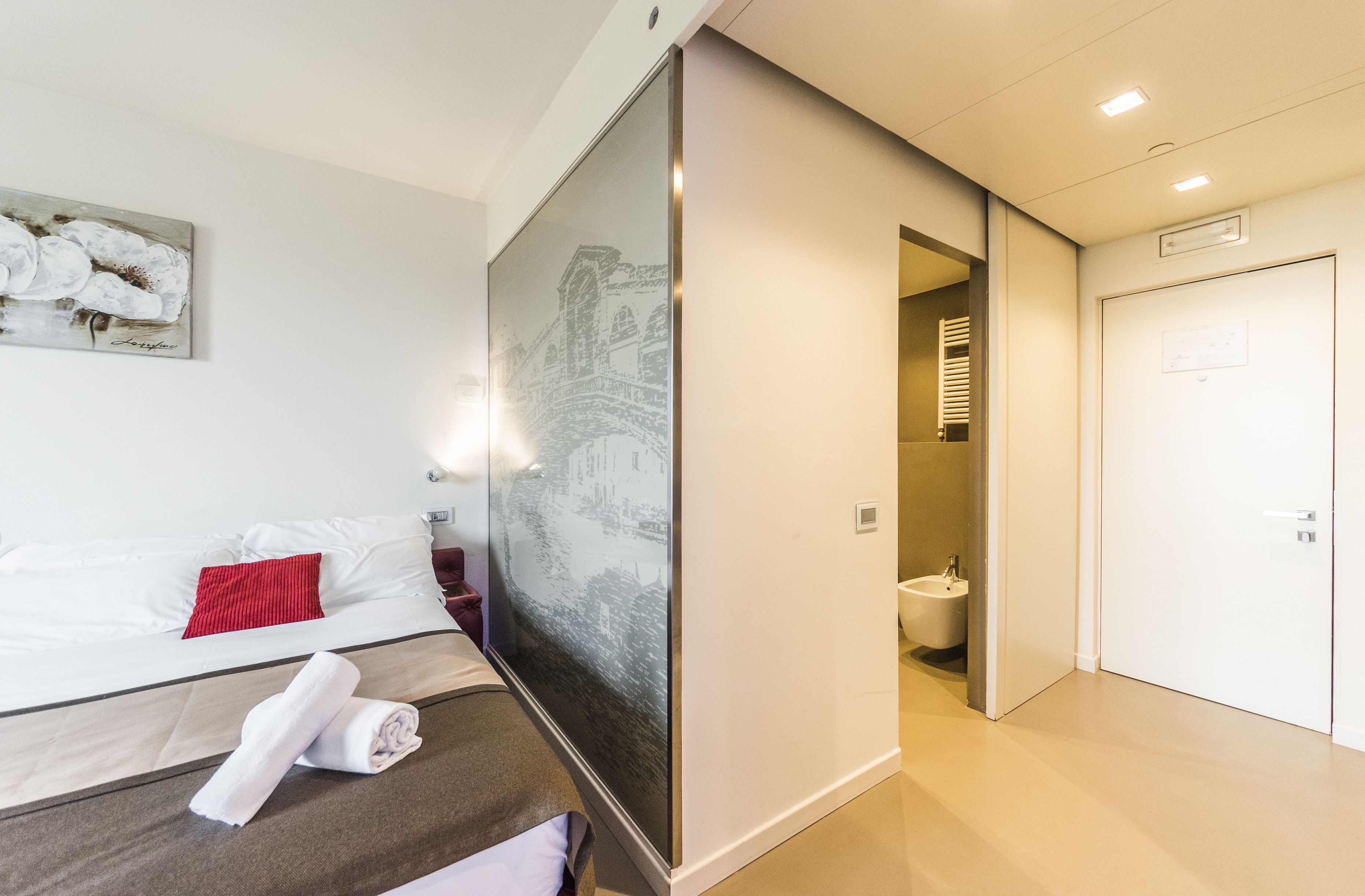 A hotel room with a bed, a painting, a bathroom, and a door.