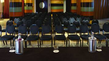 Une salle de conférence avec des rangées de chaises, un pupitre et une table équipée d’un microphone et de documents.