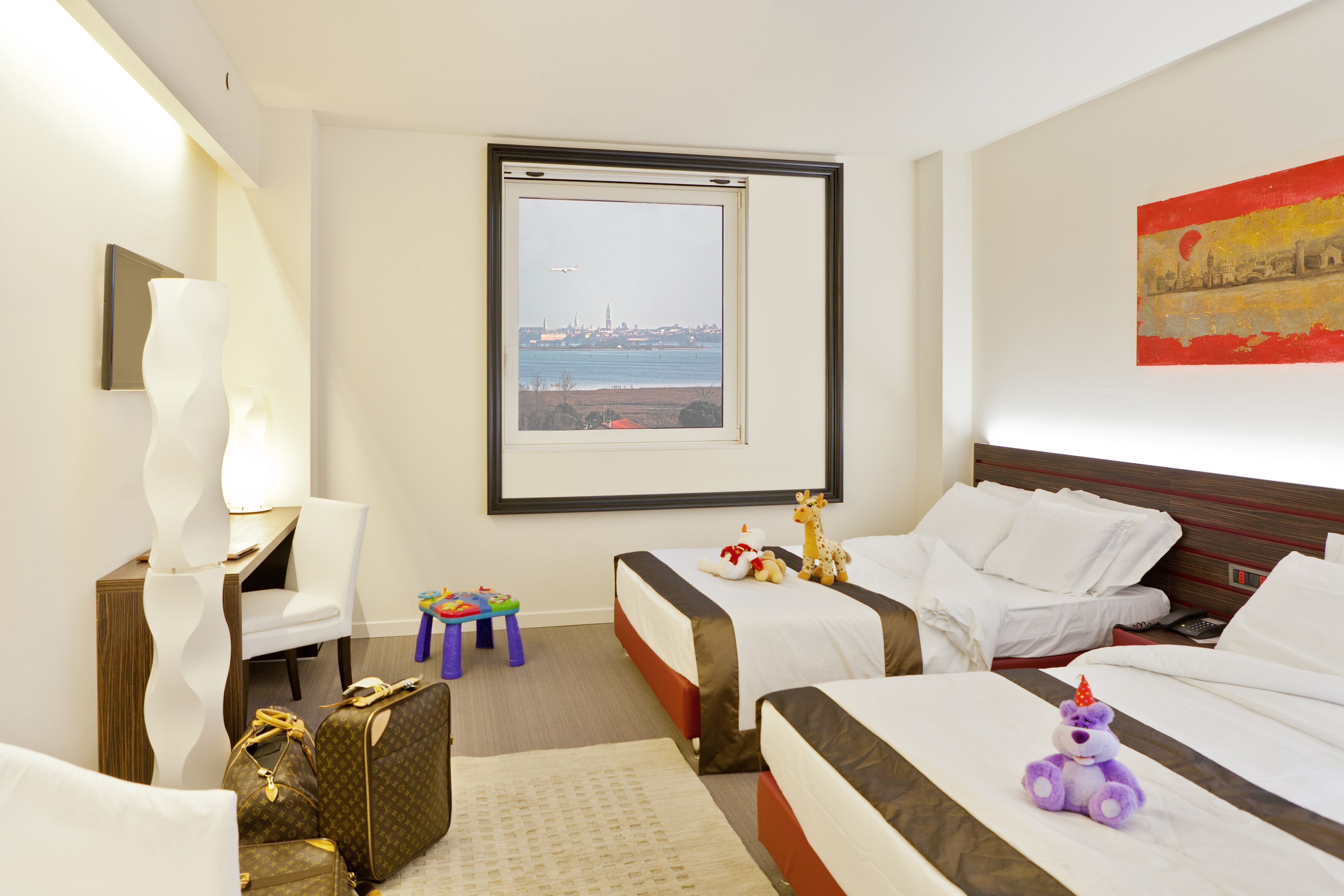 Ett hotellrum med två sängar, ett skrivbord, en stol, ett fönster med utsikt över staden och ett litet bord med leksaker.