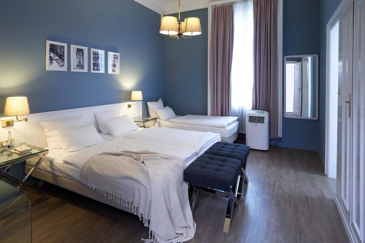Une chambre d’hôtel avec deux lits, un banc, une table de chevet avec des lampes, un radiateur et un miroir au mur.