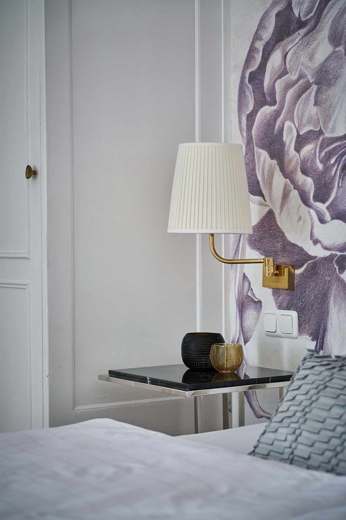 A neatly made bed with a white comforter, a black bedside table with a gold lamp, and a wall with a purple floral pattern.