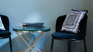 A room with a blue textured wall, a framed picture of a chandelier, a round glass table with metal legs, and two dark chairs with a patterned pillow.