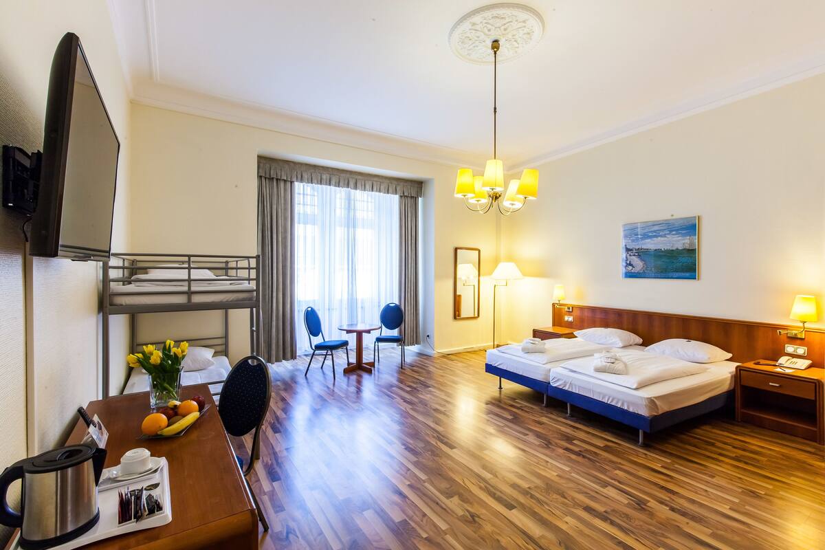 Une chambre d’hôtel avec un lit superposé, un bureau avec une bouilloire, une chaise, une table avec des fruits et un tableau accroché au mur.