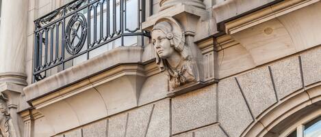 A building facade with a classical architectural style, featuring a detailed stone relief sculpture, arched windows, and ornate iron balcony railings.