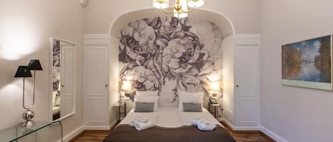 A bedroom with a large bed, a chandelier, a floral mural, a painting, and a glass table.