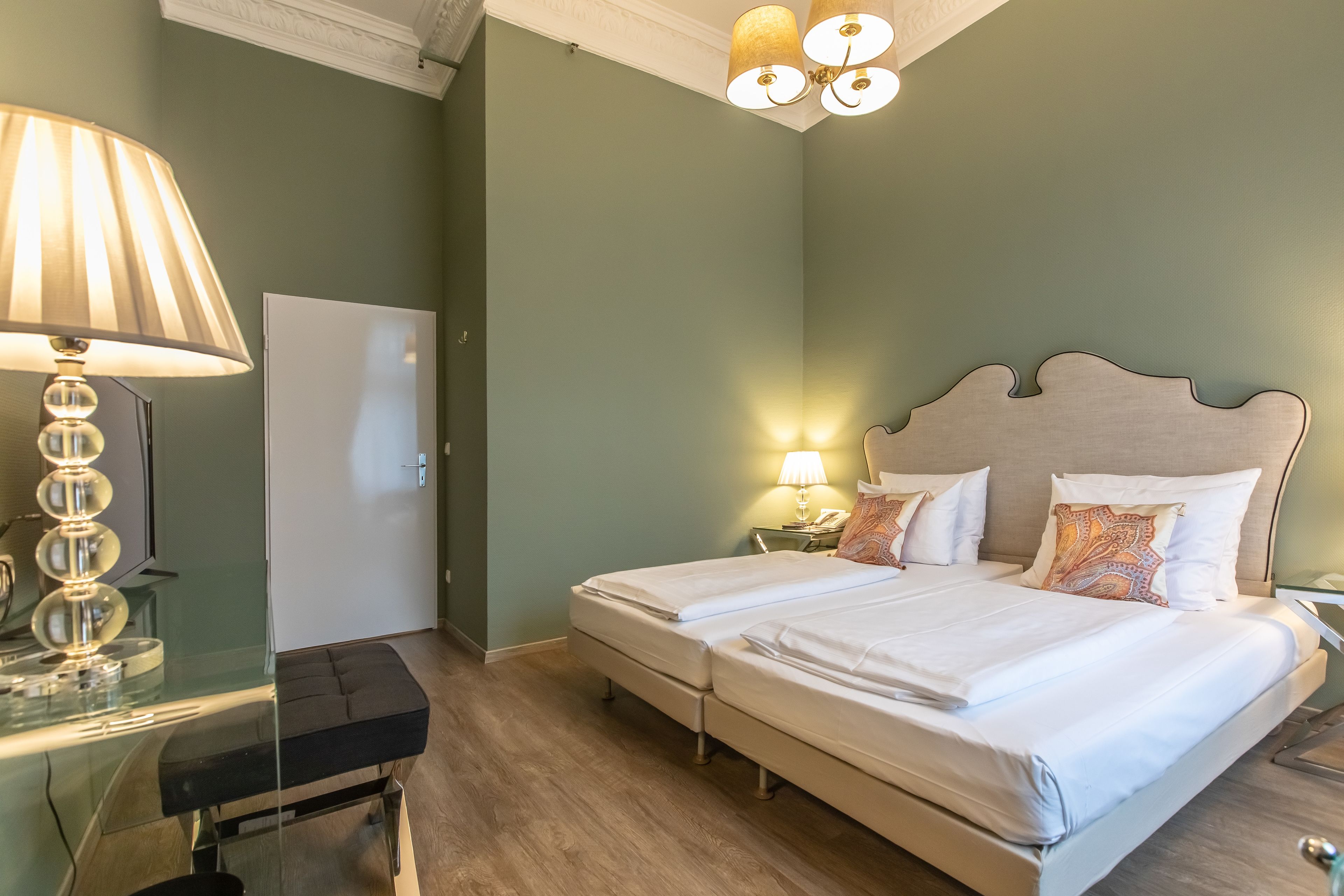 A double bed with a headboard, bedside tables with lamps, a bench, and a wardrobe in a room with green walls and wooden flooring.