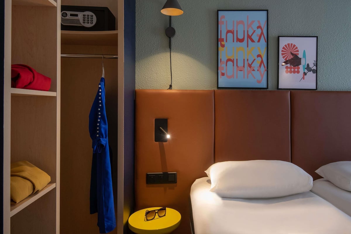 A hotel room with a bed, a bedside table with glasses, a coat rack, a wardrobe, and two framed pictures on the wall.