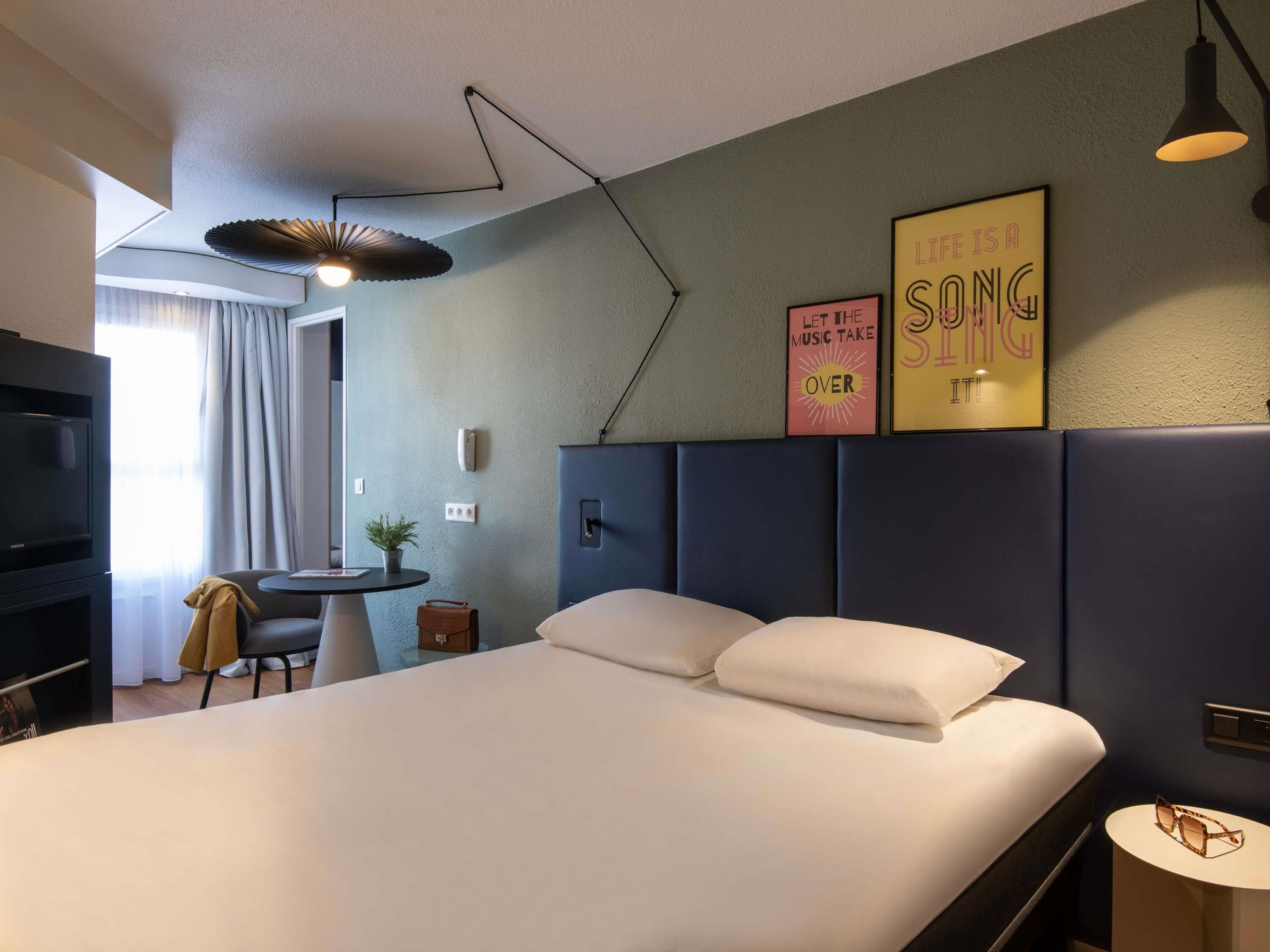 A modern hotel room with a large bed, a desk, a television, and a wall-mounted light fixture.