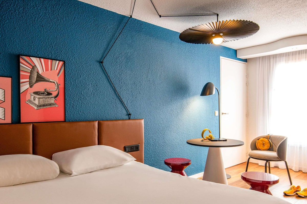 A modern hotel room with a large bed, a small round table, a chair, and a unique ceiling light fixture.