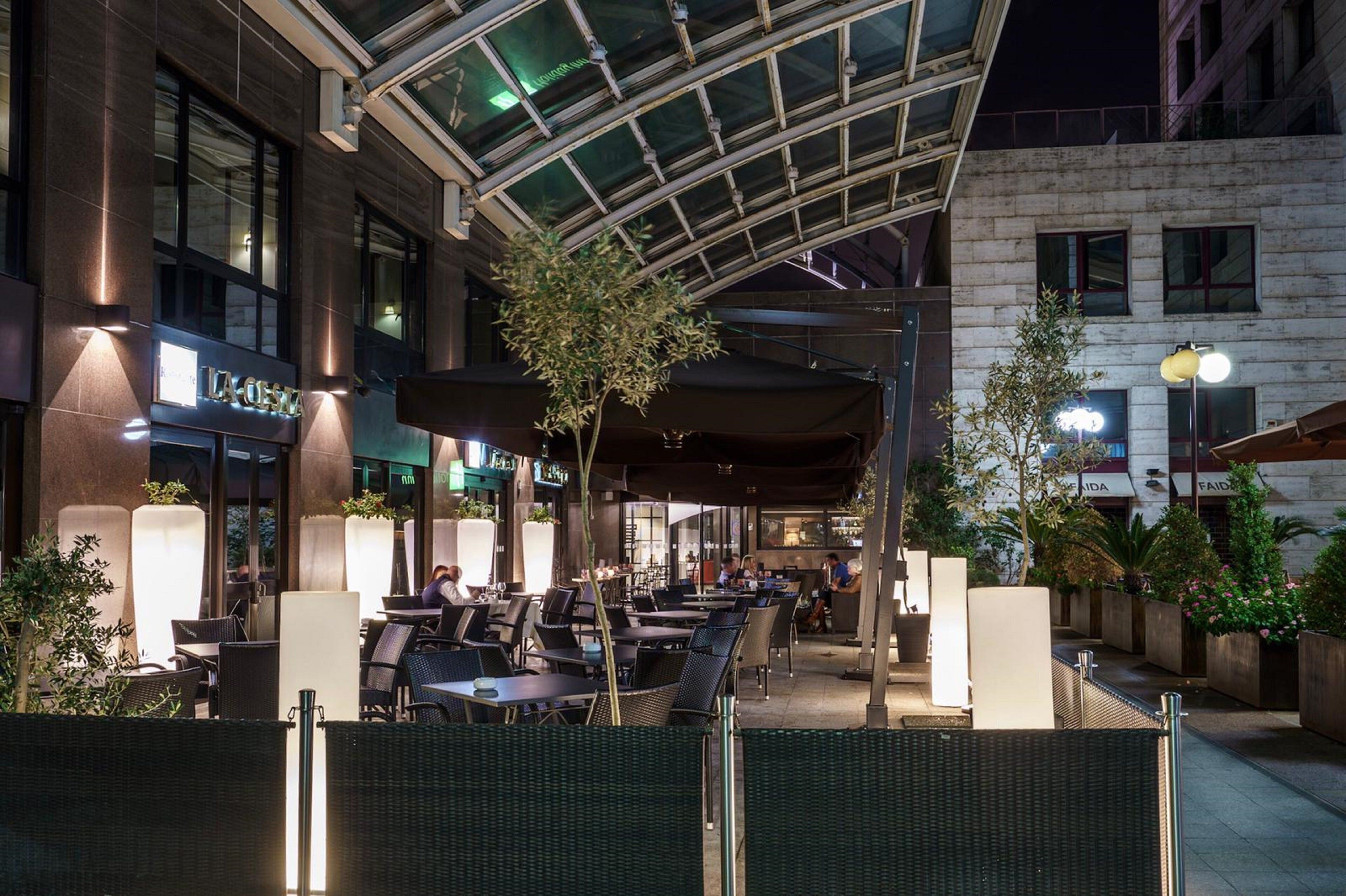 An outdoor cafe area with tables and chairs, a glass ceiling structure, and a building with a sign that reads "LA CASA".