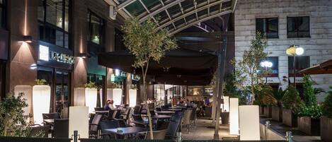 An outdoor cafe area with tables and chairs, a glass ceiling structure, and a building with a sign that reads "LA CASA".