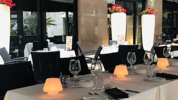 A restaurant with tables set for dining, featuring white tablecloths, black chairs, and candlelit lamps.