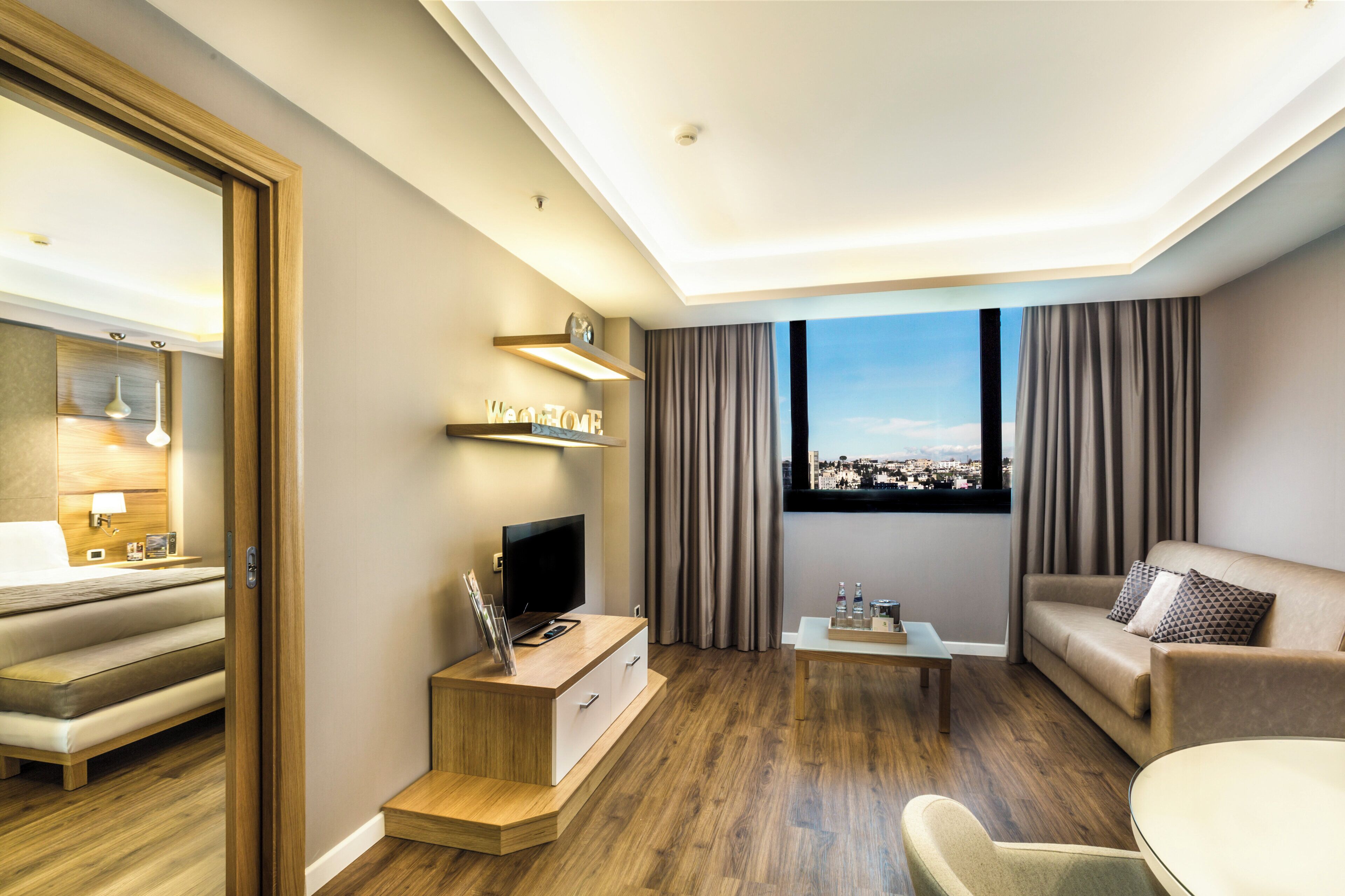 Una habitación de hotel moderna con un televisor de pantalla plana, un sofá, una mesa de centro y vistas a un paisaje urbano a través del ventanal.