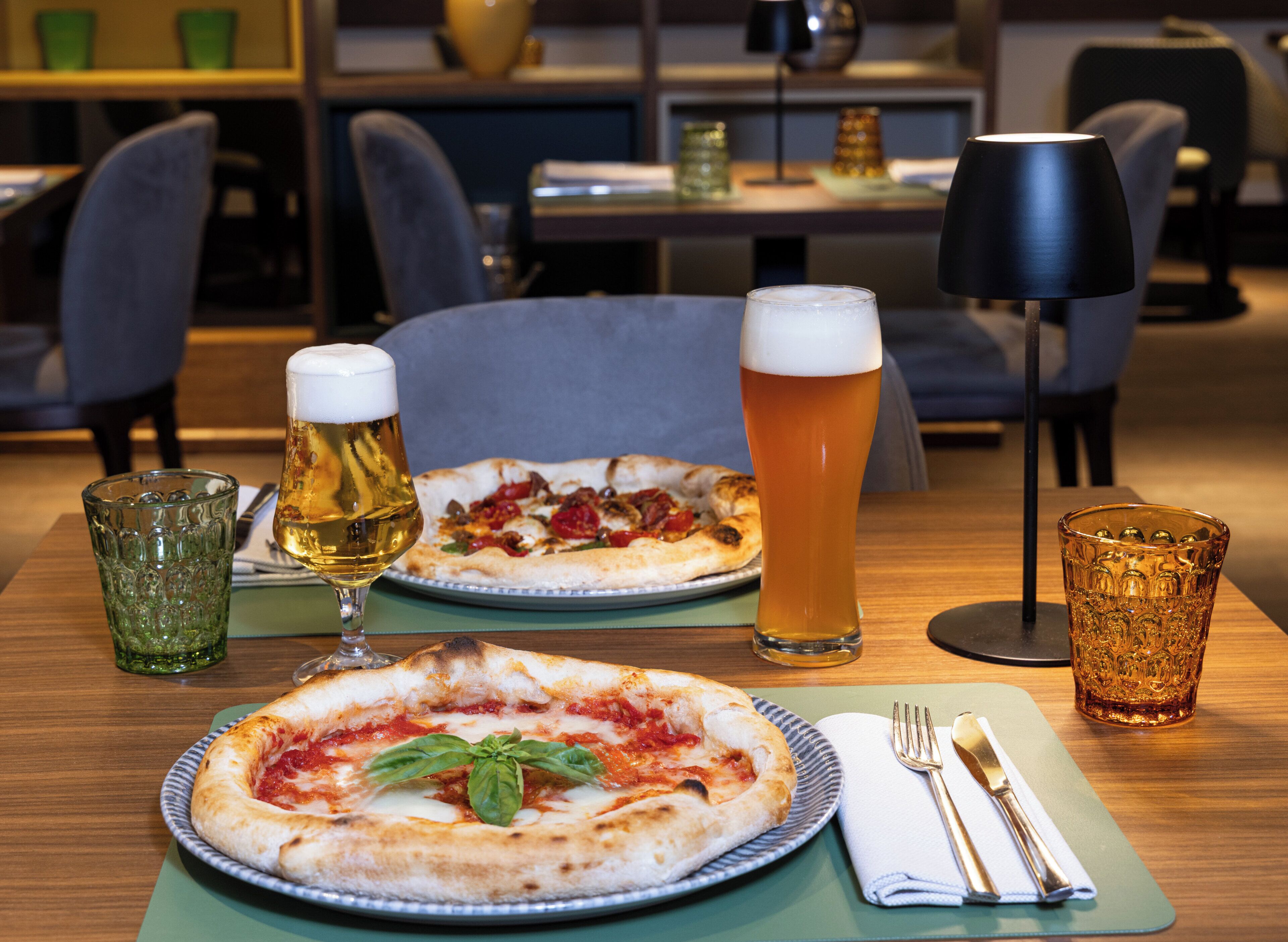Une table à manger avec deux pizzas, deux verres de bière et une lampe.