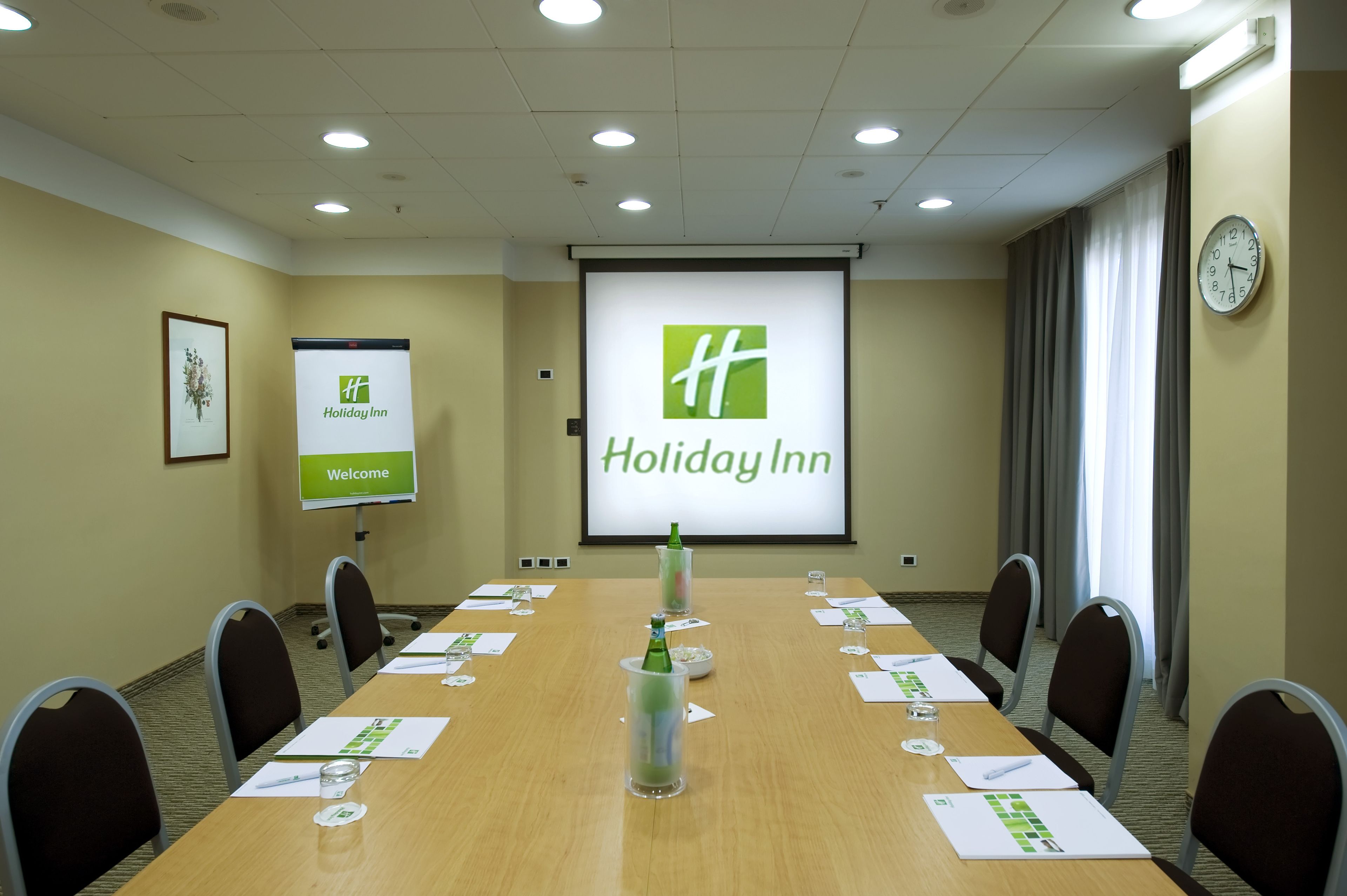 A conference room with a long table, chairs, a projector screen displaying 'Holiday Inn', and a clock on the wall.