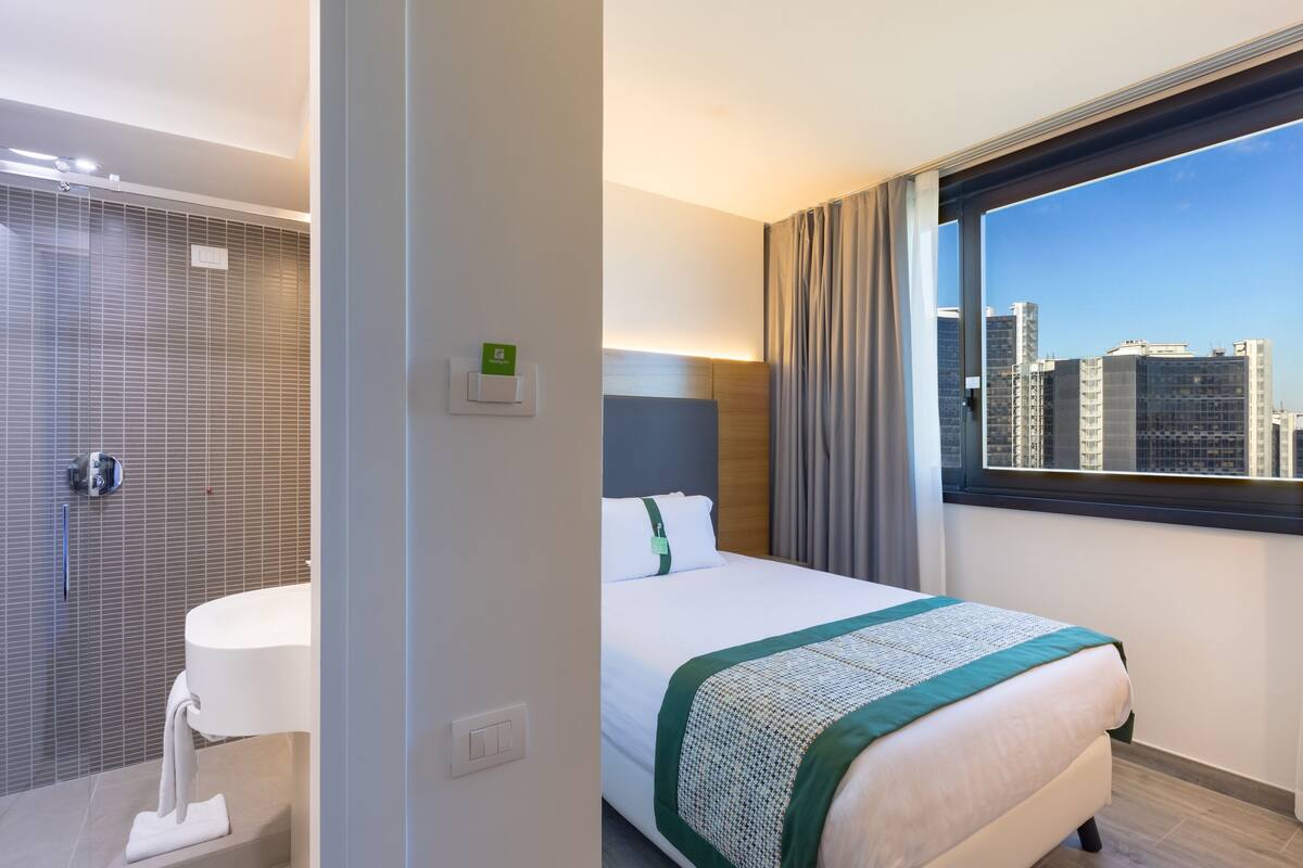 A hotel room with a bed, a bathroom with a sink, and a view of the city skyline through a window.