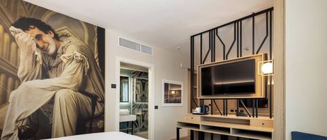 Quarto de hotel com uma cama grande, televisão fixada num painel de madeira, poltrona azul e parede com uma pintura grande.
