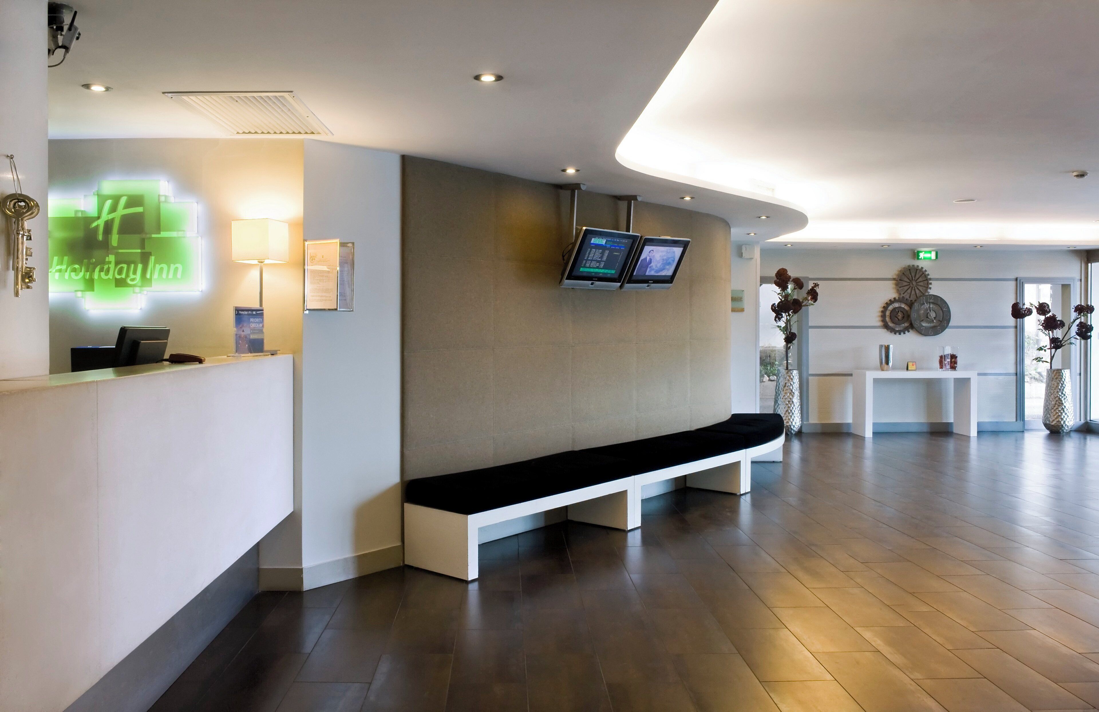 Ein moderner Hotel-Empfangsbereich mit einer Rezeption, digitalen Bildschirmen, einer Bank und einer Wanduhr.