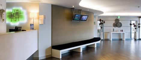 A modern hotel reception area with a reception desk, digital screens, a bench, and a clock on the wall.