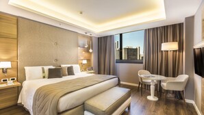 Ett modernt hotellrum med en stor säng, sängbord, en sittgrupp med bord och stolar samt utsikt över staden genom fönstret.