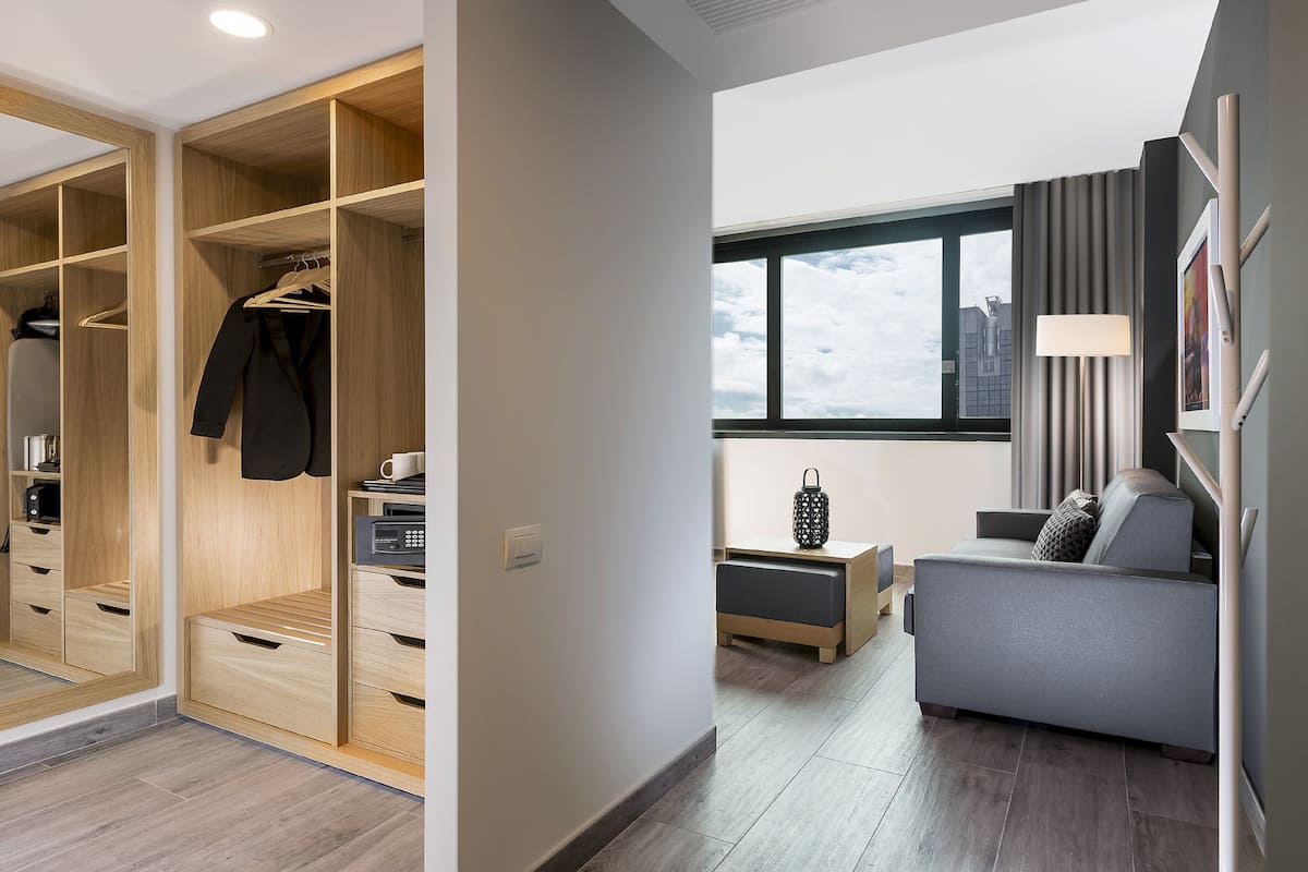 Une chambre moderne avec un canapé gris, une armoire en bois et une grande fenêtre donnant sur le ciel.