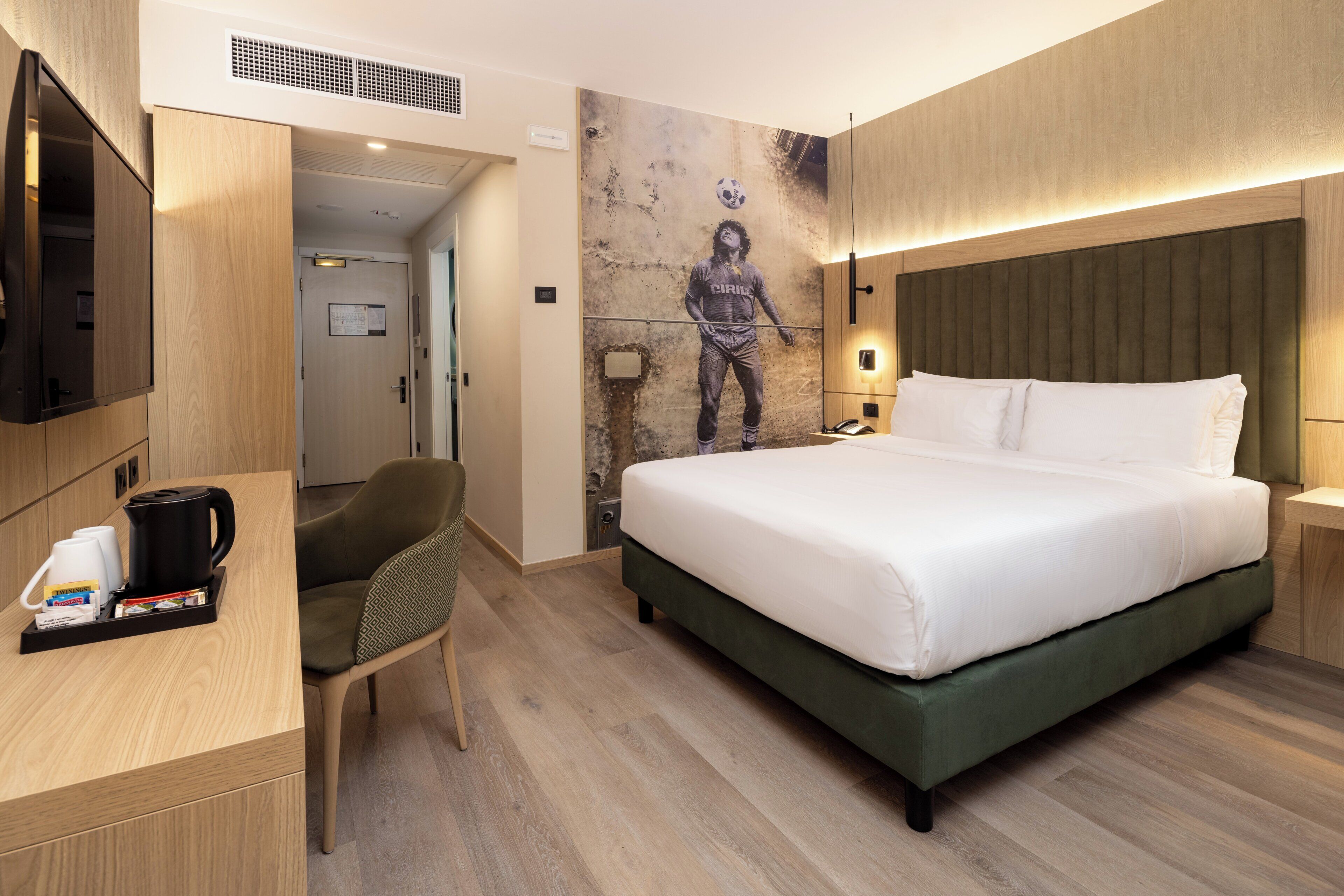 A hotel room with a bed, a desk with a coffee maker, a chair, a wall-mounted TV, and a mural.