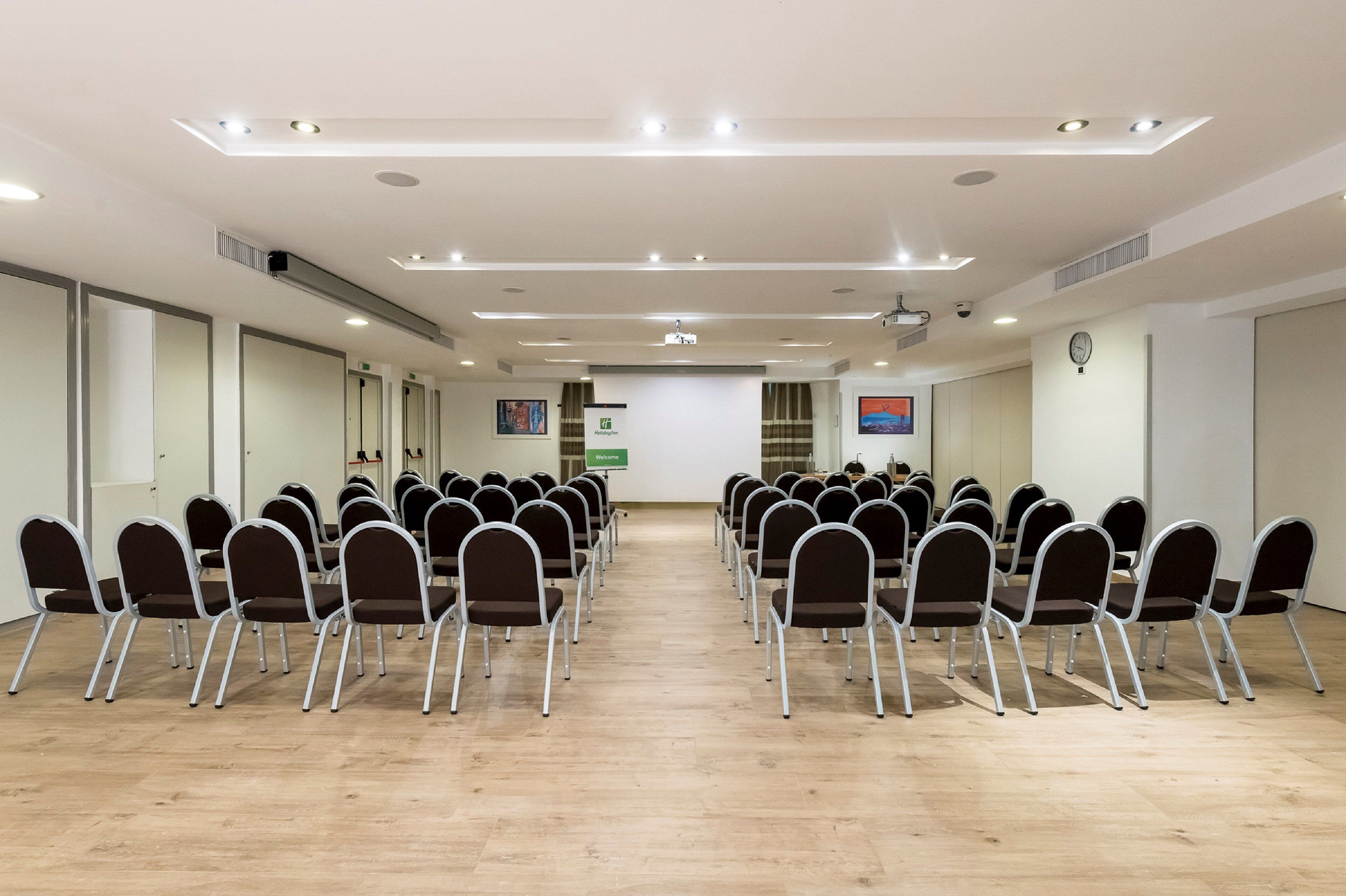 Ein Konferenzraum mit Reihen aus klappstühlen, einer Projektionsleinwand und einer Uhr an der Wand.
