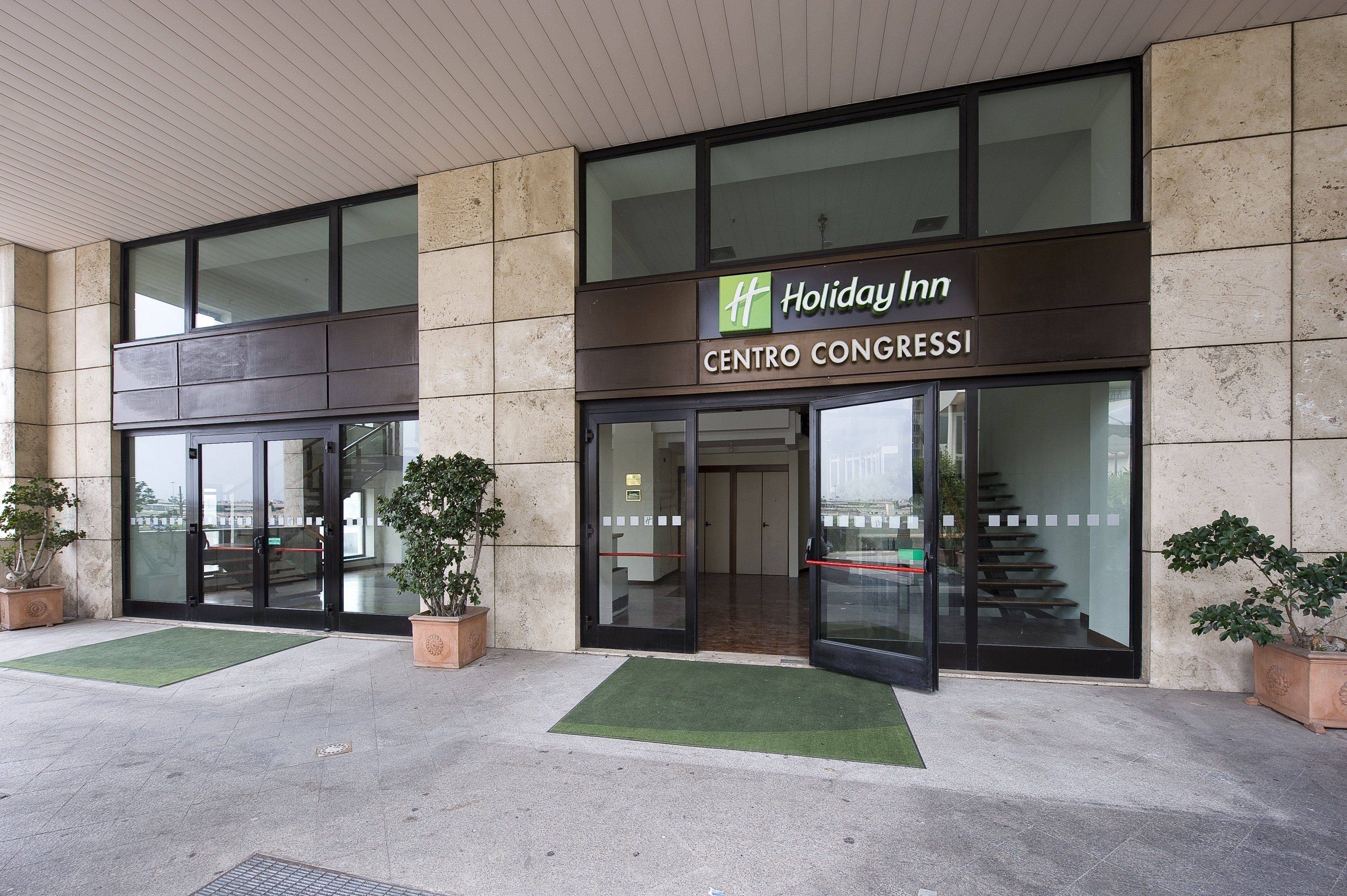 The exterior of Holiday Inn Centro Congressi with a modern design, large glass windows, and a green entrance mat.