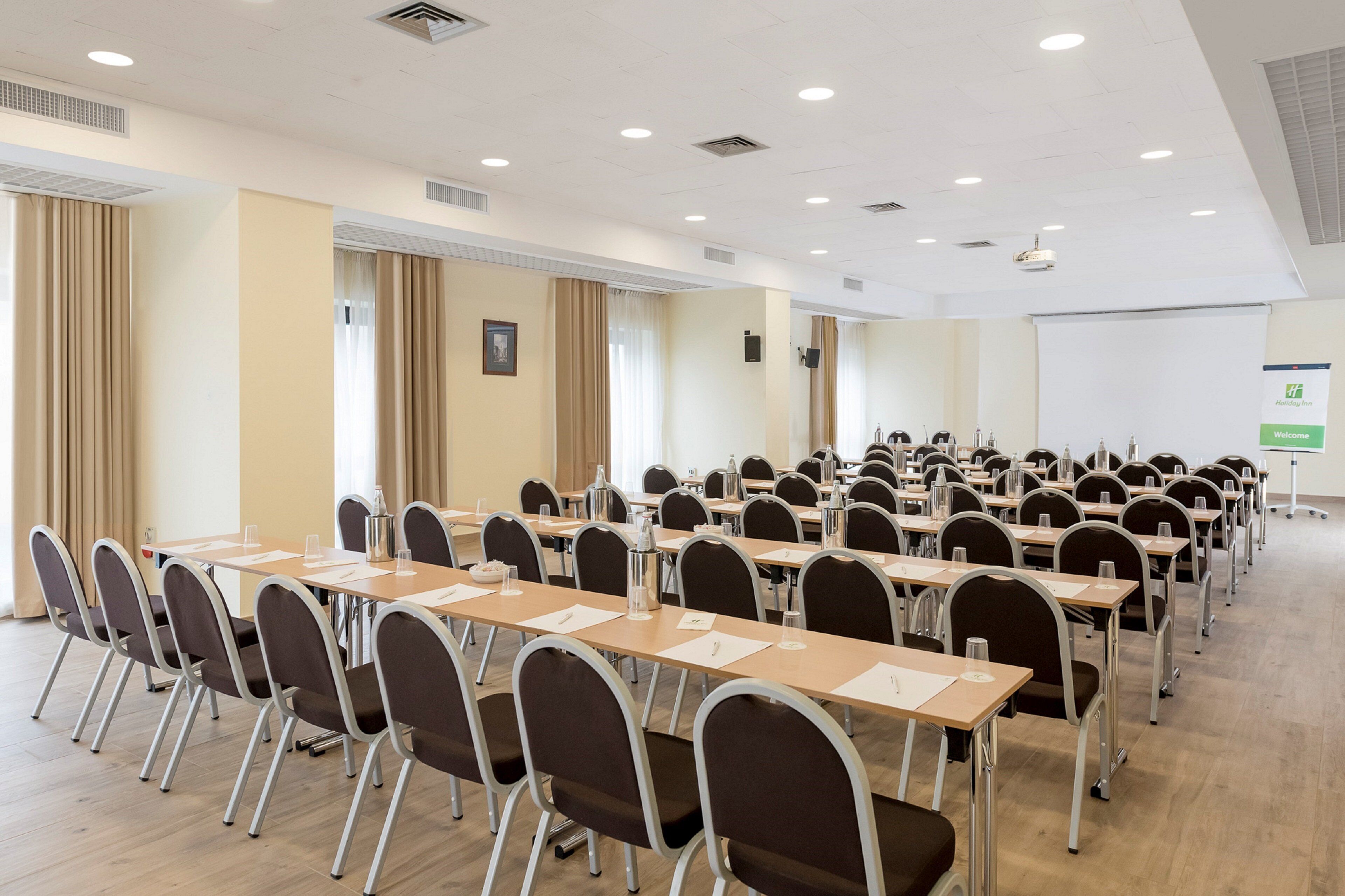 Uma sala de conferências com fileiras de mesas e cadeiras, uma tela de projetor e um letreiro com a palavra 'Welcome'.