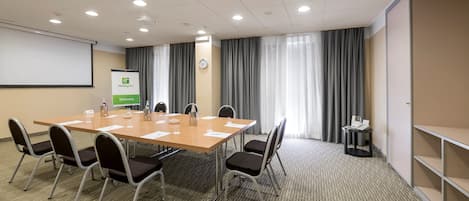 Un salón de conferencias con una larga mesa, sillas, una pantalla de proyector, un reloj y un cartel.