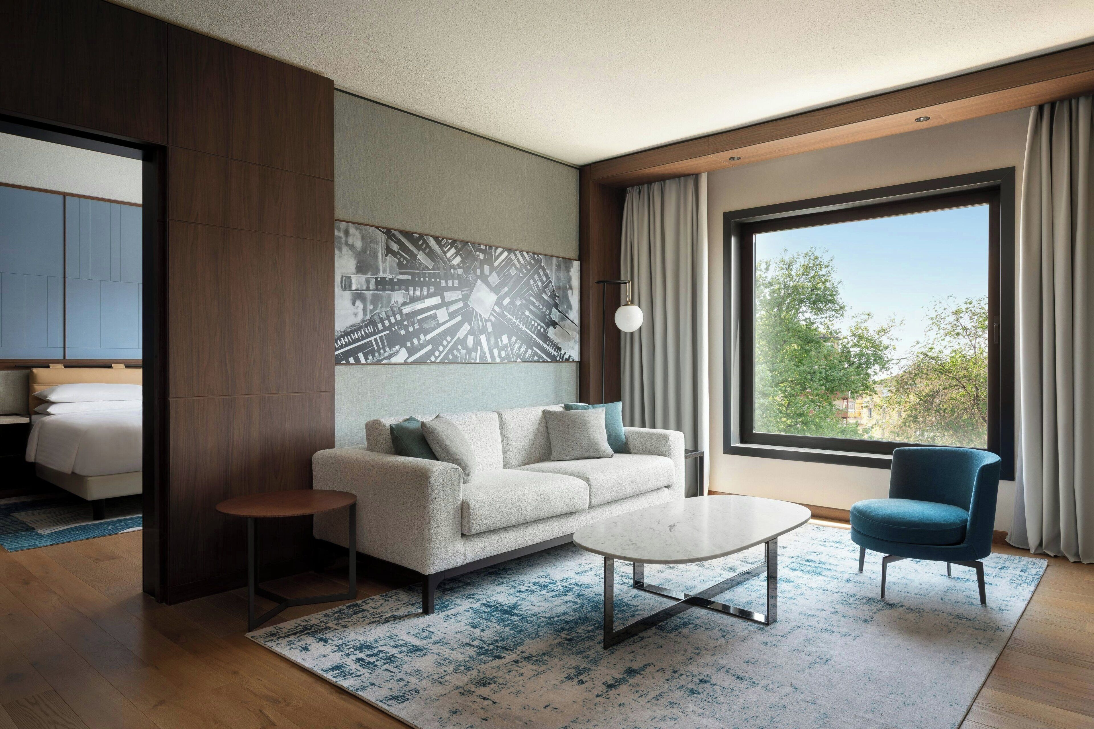 A modern hotel room with a sofa, a coffee table, a blue armchair, a large window with a view, and a painting on the wall.