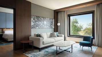 A modern hotel room with a sofa, a coffee table, a blue armchair, a large window with a view, and a painting on the wall.